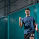 Egy fiatal férfi boldogan ünnepel a sportcsarnokban, teniszütővel a kezében, motiváltan a sport iránt.