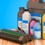Toner és tintapatronok különböző színekben, egy asztalon elhelyezve, háttérben kék színnel.