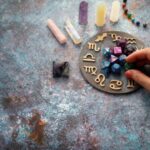 Egy kéz asztalon tarot kockákat helyez el egy asztrológiai táblán, körülötte különböző kristályok és eszközök találhatók.