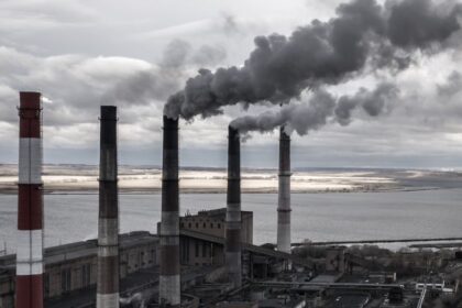 Fosszilis energia termelését bemutató ipari táj, füstölgő kéményekkel és vízparttal.