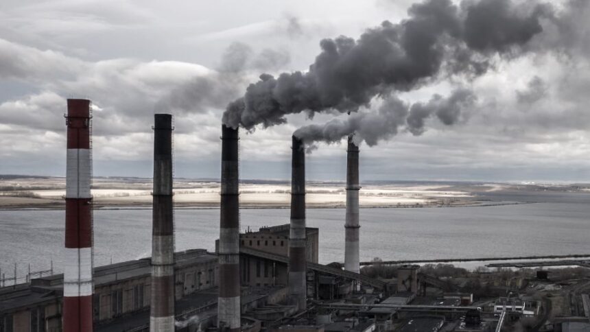 Fosszilis energia termelését bemutató ipari táj, füstölgő kéményekkel és vízparttal.