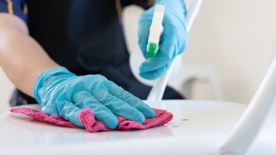 Egy kéz kék gumikesztyűben, pink törlőkendővel takarít egy fehér széket.
