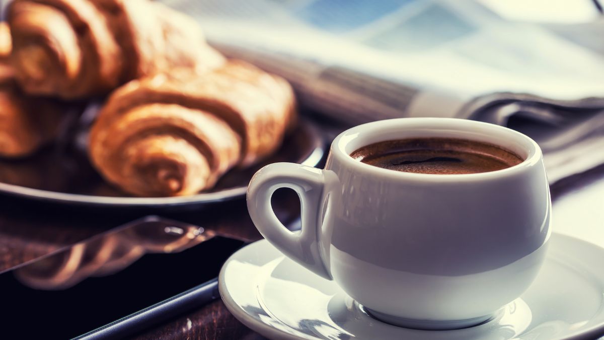 Egy csésze friss kávé áll egy asztalon, mellette croissant és egy telefon.