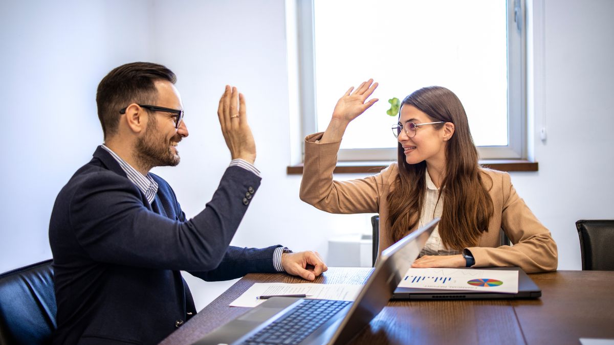 Két üzletember, férfi és nő, vidáman integetnek egymásnak egy irodában, laptop és dokumentumok láthatók az asztalon.