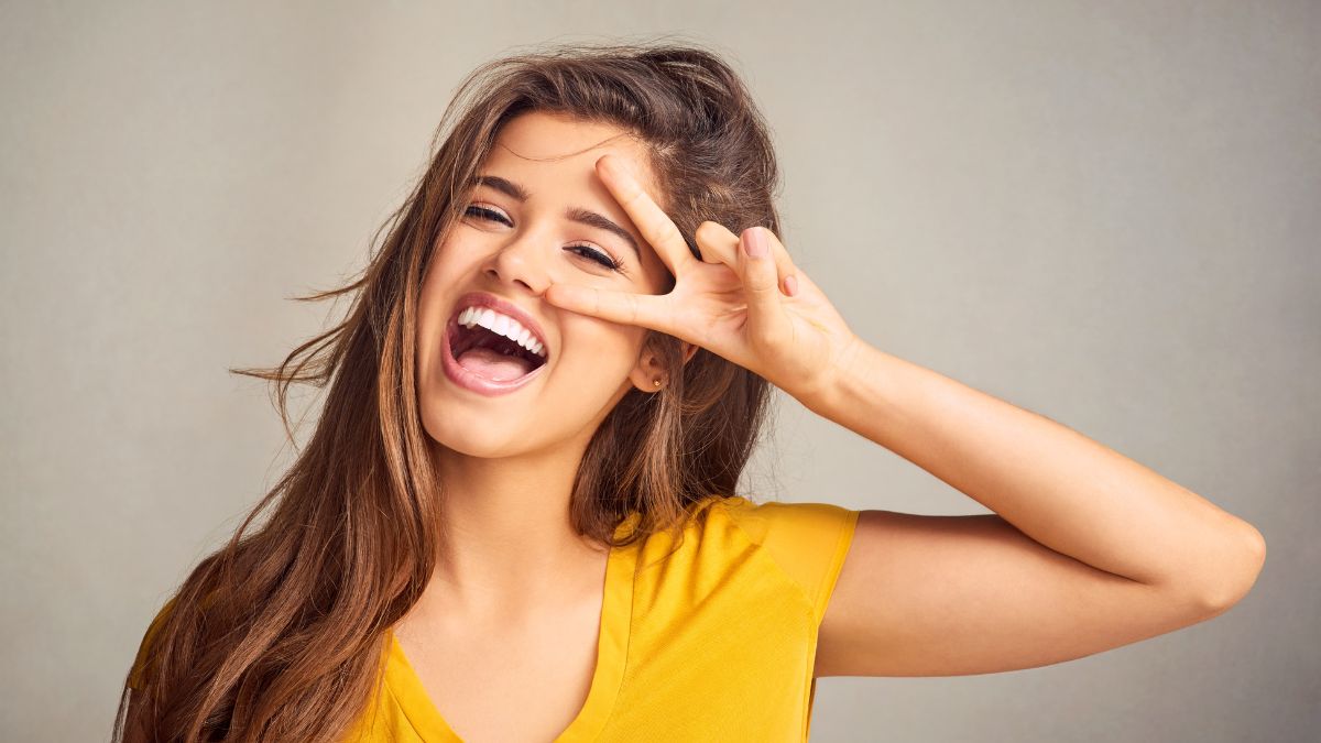 Egy fiatal nő széles mosollyal és vidám arckifejezéssel, sárga pólóban, a kezével békét mutatva.